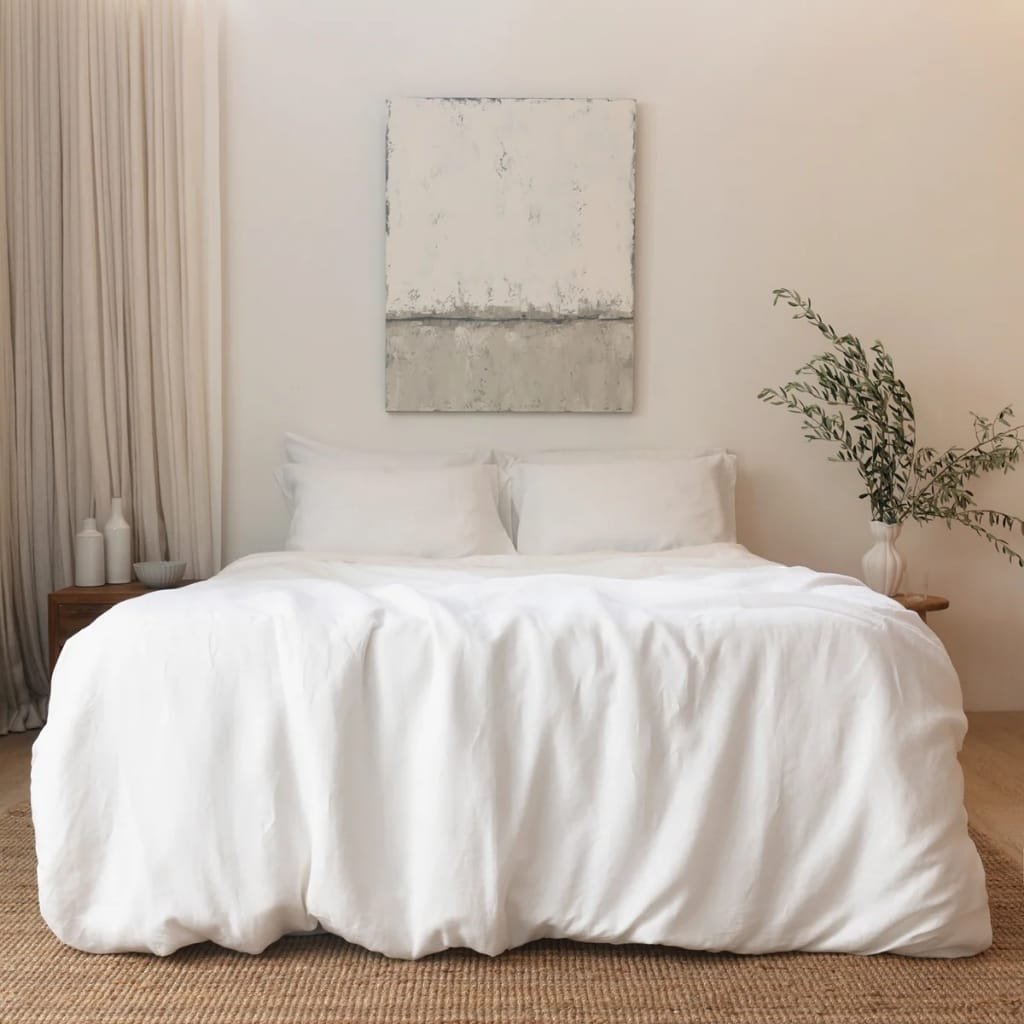 A neatly made bed draped in white linens with two pillows, positioned against a minimalist white wall. A piece of abstract art hangs above the bed, and to the right, a vase with green foliage sits on a small wooden side table. To the left, off-white curtains cover a window.