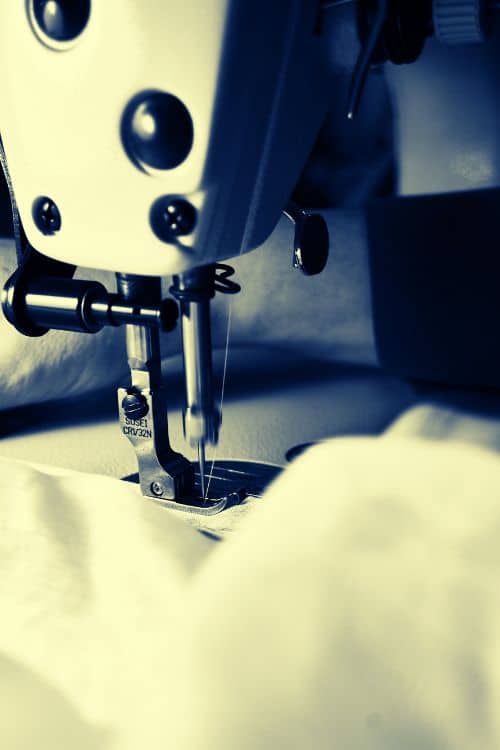 A close-up view of a sewing machine needle and presser foot, with fabric underneath, captured in monochrome tones.