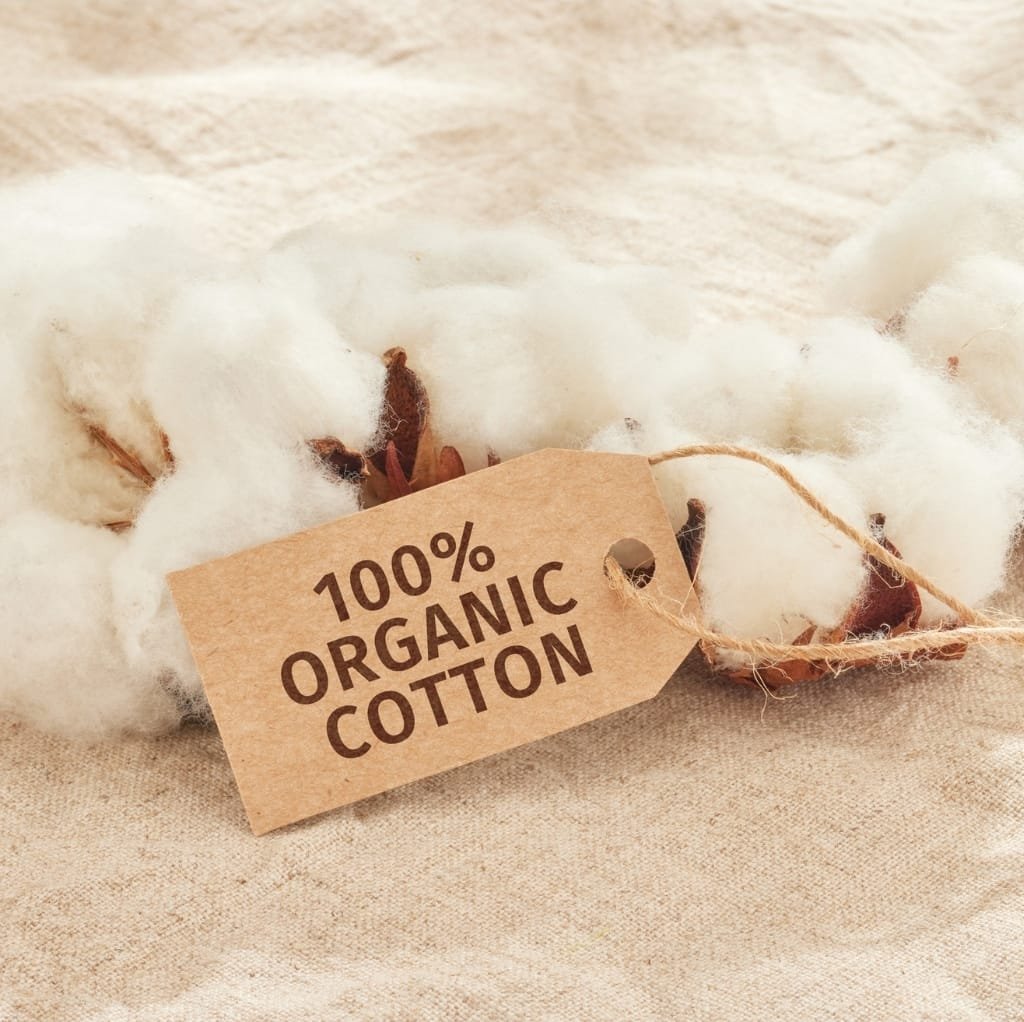 A close-up of a tag attached to a piece of white cotton, with a background of soft, natural fabric. The tag reads "100% ORGANIC COTTON."