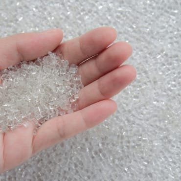 A person's hand holding a handful of small, clear, cube-shaped plastic PET chips, with a large pile of similar chips spread out on a surface in the background.