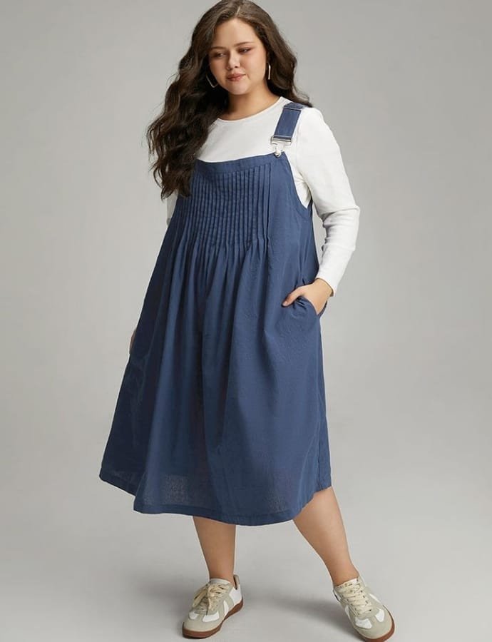 A plus-size woman with long, wavy brown hair wearing a casual denim pinafore dress over a white long-sleeve top, paired with beige sneakers, standing with one hand in her pocket against a neutral background.