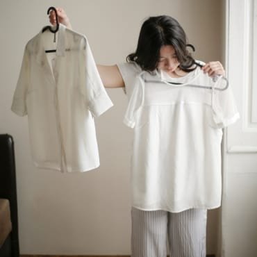 A woman holding up two white polyester shirts, one in each hand, indoors near a window. She is wearing striped pajamas and looking at the shirts with interest.