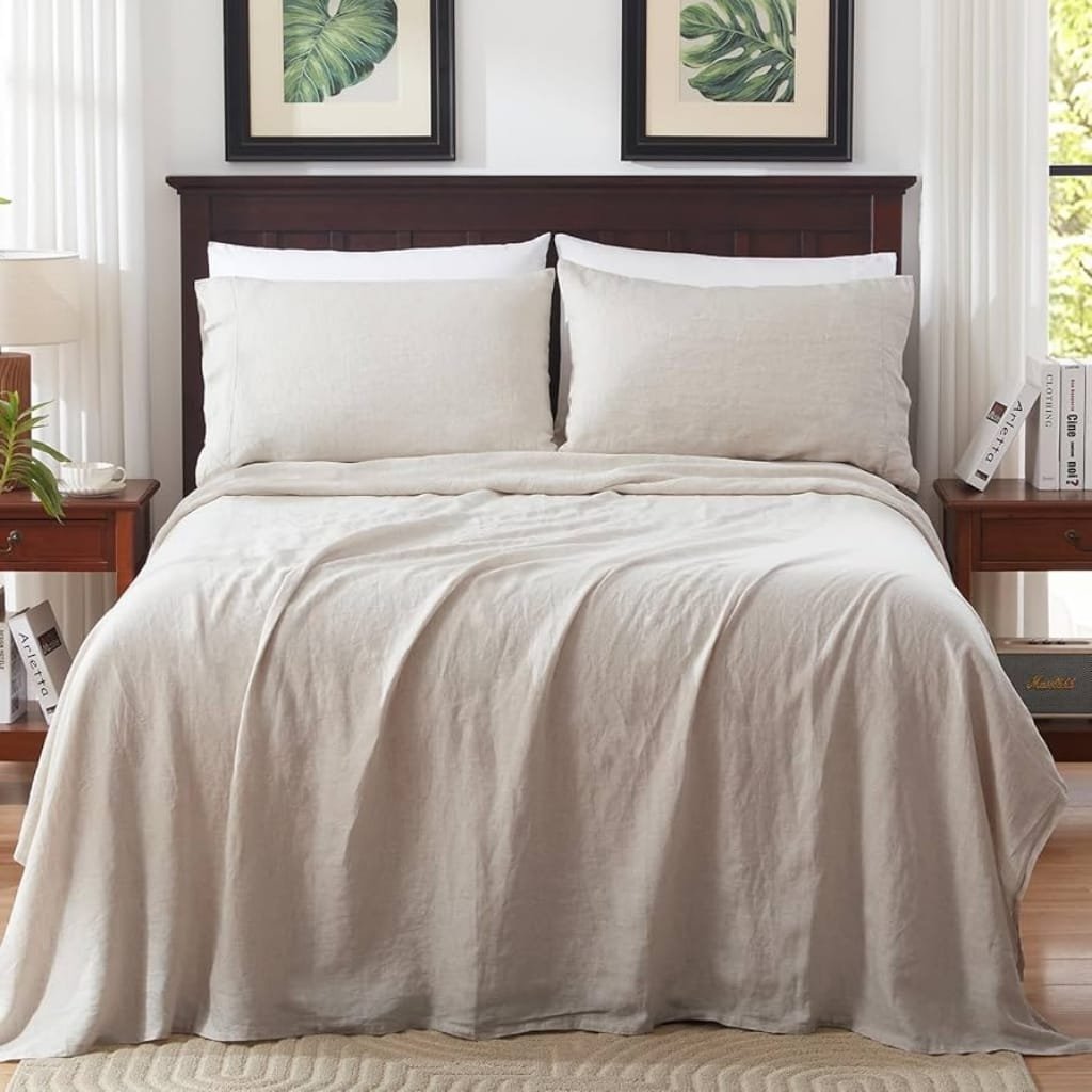 A neatly made bed with a dark wooden headboard, covered in a light cream or beige bedspread, with two matching pillows. There are two nightstands on either side of the bed, each holding books, a small plant, and a lamp. Two framed botanical prints hang above the headboard, and light filters in through white curtains on the windows beside the bed.