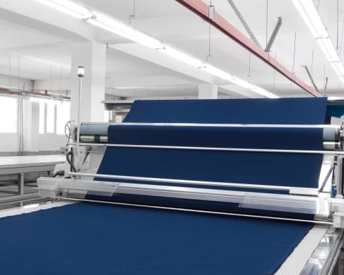 A large industrial fabric processing machine in a clean, modern factory, with navy blue fabric being fed through the rollers.