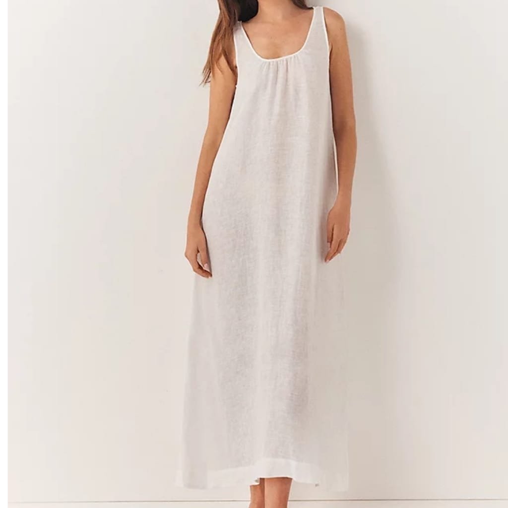 A woman wearing a long, sleeveless, light-colored summer dress with a round neckline, standing against a plain wall.