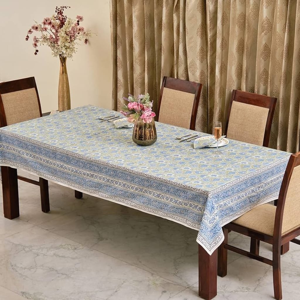 A dining table covered with a blue and white patterned tablecloth, set with neatly folded cloth napkins, utensils, and a glass with a pink beverage. There are two floral arrangements: one in a tall gold vase in the background with pink and white flowers, and a smaller one in a dark vase with pink and purple flowers in the center of the table. Four wooden chairs with beige cushions are positioned around the table, and beige patterned curtains hang in the background.