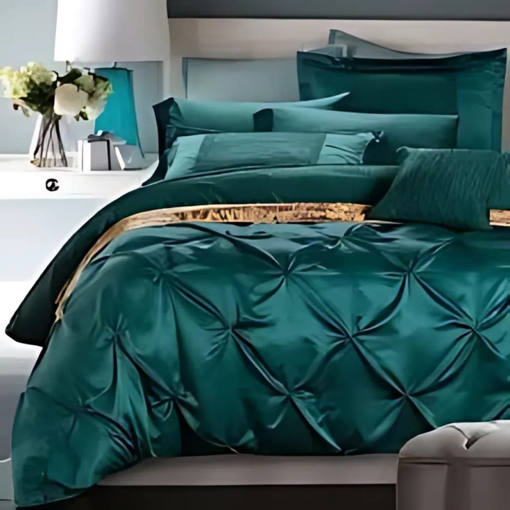 A neatly made bed with emerald green satin bedding, featuring a tufted comforter and matching pillows. There is a white nightstand beside the bed with a contemporary lamp, a small vase of white flowers, and a decorative object. The room has a modern and elegant decor style.
