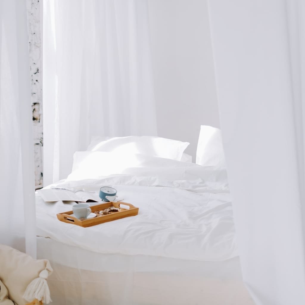 A bright, minimalist bedroom with white walls, white bed linens, and sheer white curtains. On the bed, there is a wooden tray with a teacup, a small plate with snacks, an open book, and a small round device. The overall ambiance is peaceful and airy.