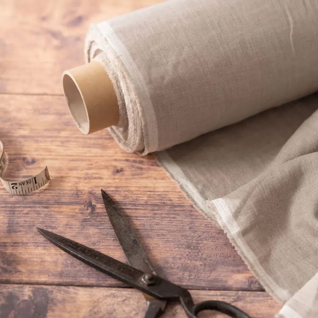 A spool of beige linen fabric on a wooden surface, with a pair of metal scissors and a measuring tape nearby.