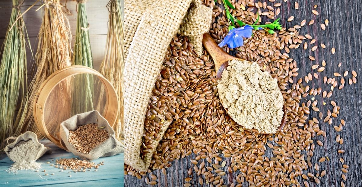 A close-up of flax seeds scattered on a dark wooden surface with a scoop of ground flaxseed, a blue flower, and a small burlap cloth. The background features a basket and bundles of dried plants hanging.