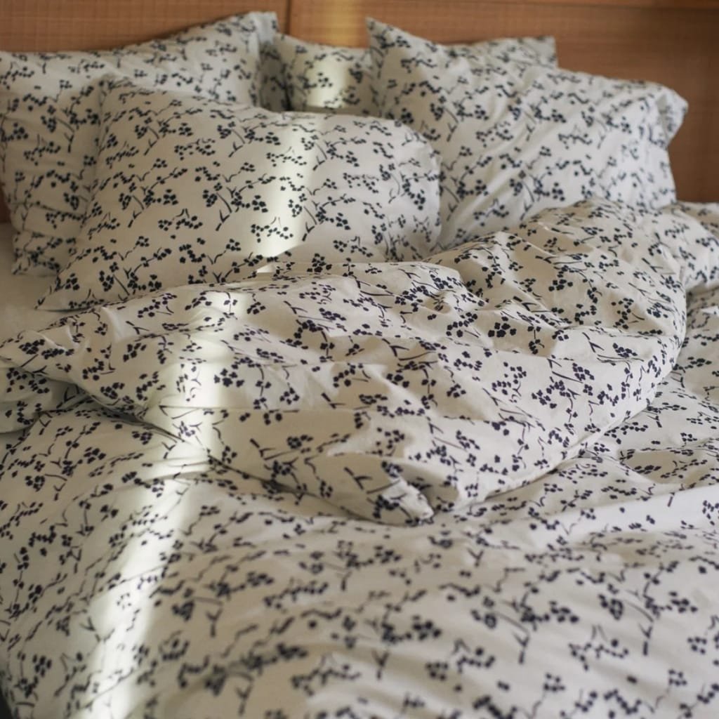 A close-up photo of a bed with white bedding decorated with a black floral or botanical pattern. The bedding appears slightly rumpled and is illuminated by natural light, creating soft shadows.