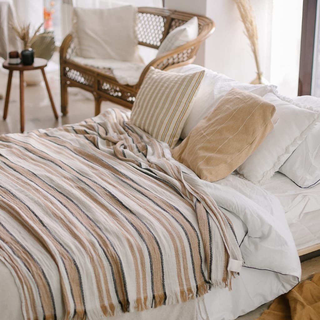 A cozy and stylish bedroom corner featuring a bed with a striped beige, cream, and black blanket, several pillows in neutral tones, and a wicker chair in the background. The room is bright with natural light coming through a nearby window