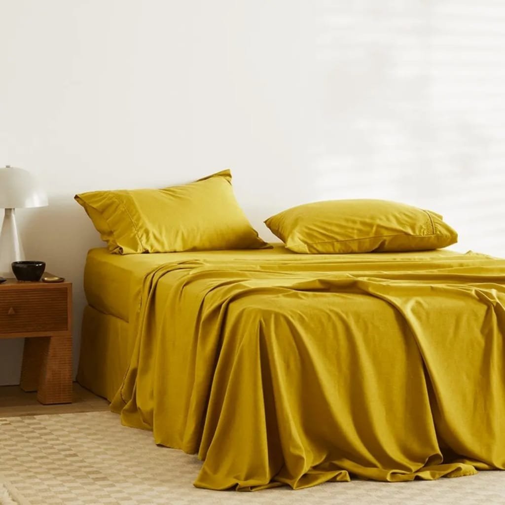 A bed with mustard yellow bedding and pillows, next to a wooden nightstand with a white lamp and a small black bowl, set against a plain white wall and a beige patterned carpet.