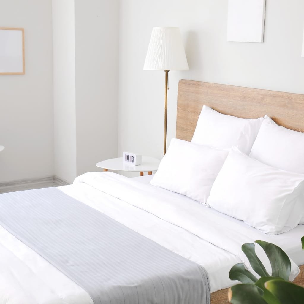 A minimalist bedroom featuring a bed with white linens and pillows, a wooden headboard, a white bedside table with a digital clock, a floor lamp, and a leafy plant in the corner.