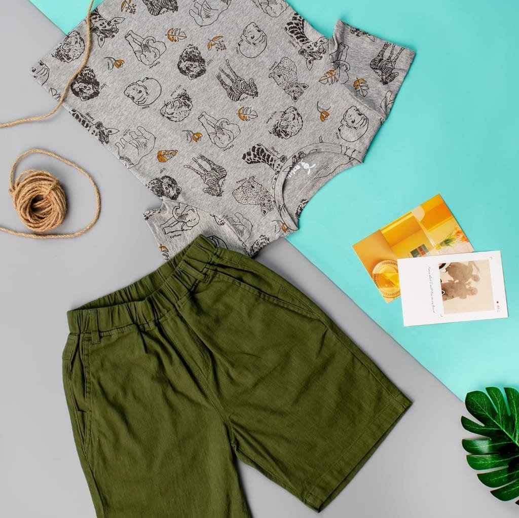 Close-up image of casual clothing items laid out on a flat surface, including a pair of green shorts, a gray t-shirt with animal illustrations, along with a ball of twine, colorful papers, and a green leaf in the background. Cotton Textiles Made in USA