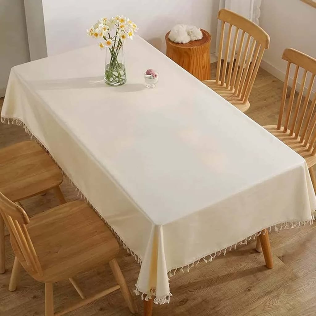 A dining table covered with a plain, light-colored tablecloth with tassel edges. The table is decorated with a vase of white flowers and a small glass container. There are four wooden chairs around the table and a wooden stool at the corner. The room has wooden flooring and a simple, cozy atmosphere.