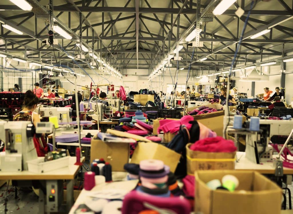 A spacious indoor clothing manufacturing facility with rows of sewing machines and workers assembling garments. The workspace is filled with colorful fabrics, spools of thread, and boxes of materials, highlighting an active garment production environment.