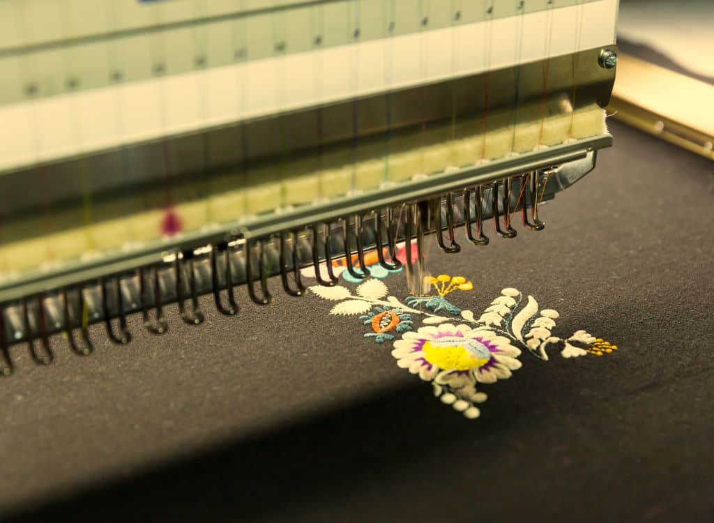Close-up of an embroidery machine stitching a colorful floral design onto dark fabric, representing clothing manufacturing in Hawaii.