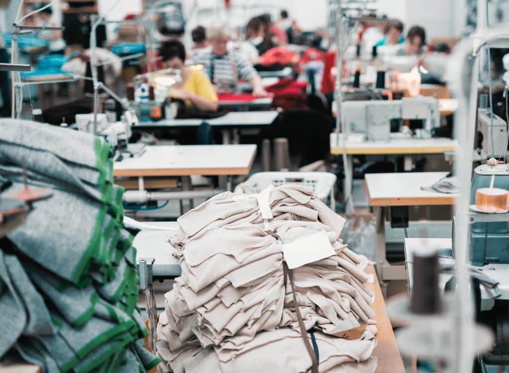 A bustling clothing manufacturing workshop with stacks of fabric and multiple sewing stations, workers in the background sewing garments.