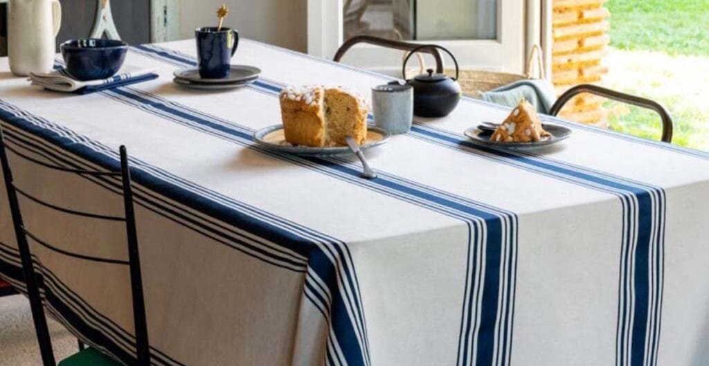 : A dining table covered with a white tablecloth featuring blue and navy stripes. The table has slices of cake on plates, a teapot, a cup, and some bowls, set near a window with a bright outdoor view.