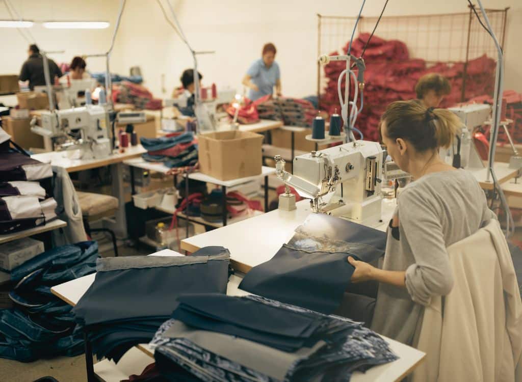 Workers operating sewing machines in a clothing manufacturing facility, surrounded by stacks of fabric and garments in progress.