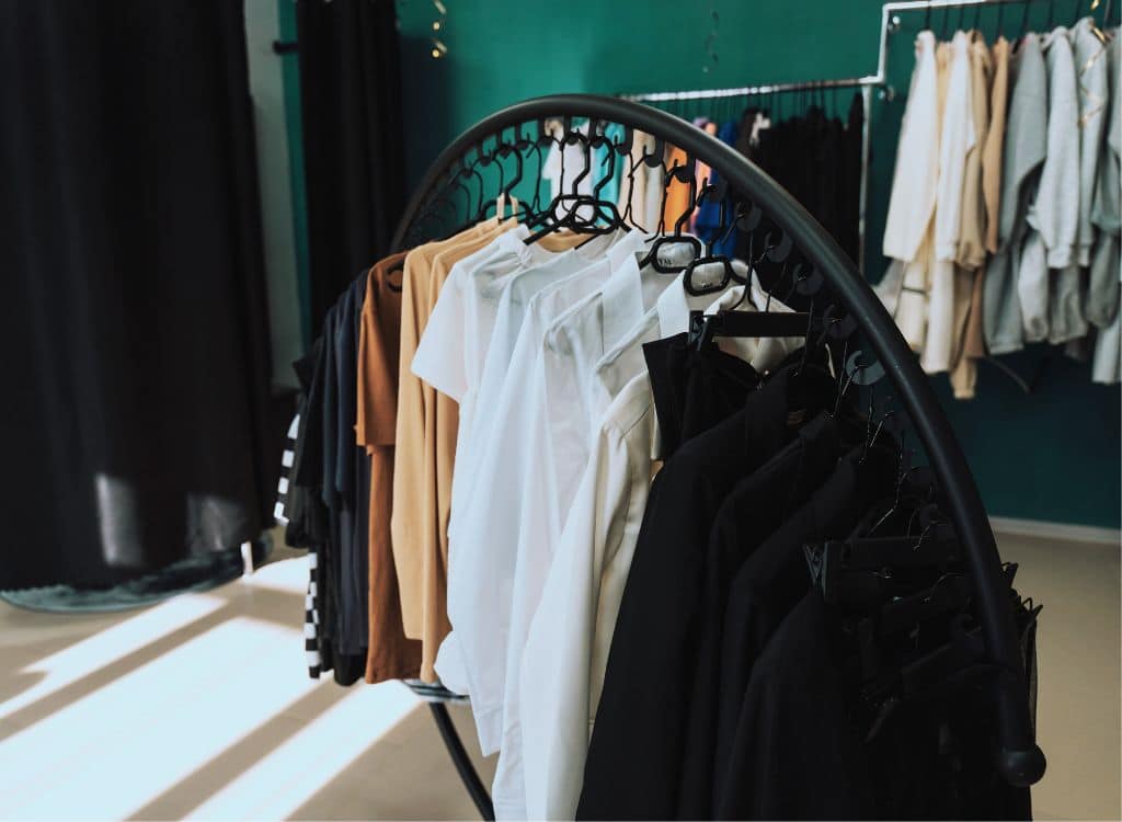 A clothing rack displaying various neutral-colored shirts and jackets inside a retail store or showroom, with a green wall and additional clothing on hangers in the background.