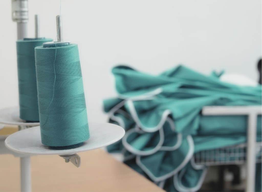 Spools of teal thread on a sewing machine with fabric in the background, inside a clothing manufacturing workshop.