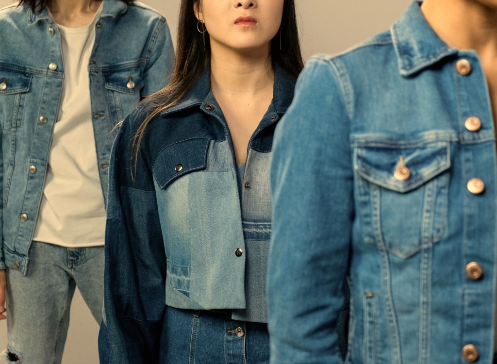 Close-up of three people wearing denim jackets, standing in a line, in a clothing manufacturing setting.