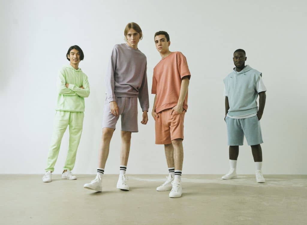 A diverse group of four young models standing in casual athletic clothing in a minimal studio setting, showcasing colorful outfits.