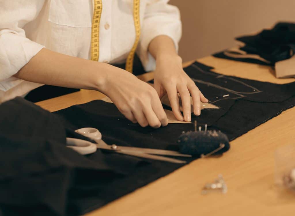 A person working on tailoring black fabric with scissors, fabric, and sewing tools on a wooden table, representing clothing manufacturing.