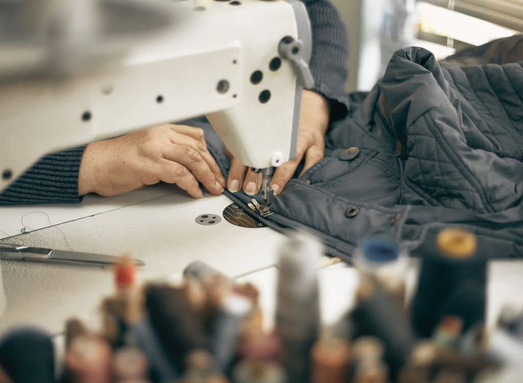 A worker sewing a jacket using a sewing machine in a clothing manufacturing workshop.