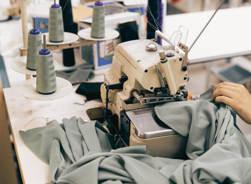Close-up of a sewing machine stitching light gray fabric, with spools of thread on a work table, representing a clothing manufacturing process.