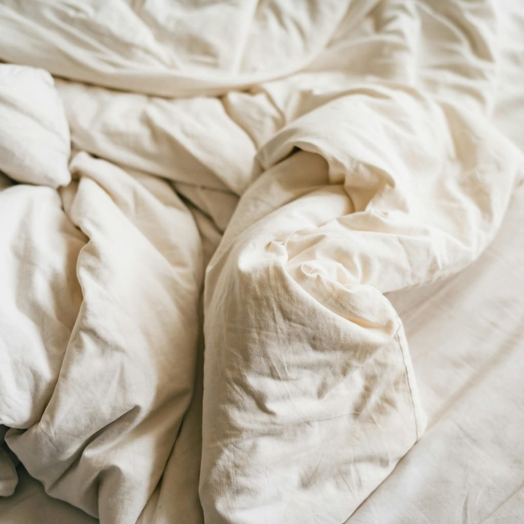 A close-up of a rumpled, cream-colored duvet cover and sheets on a bed, with soft, natural lighting highlighting the fabric's texture.