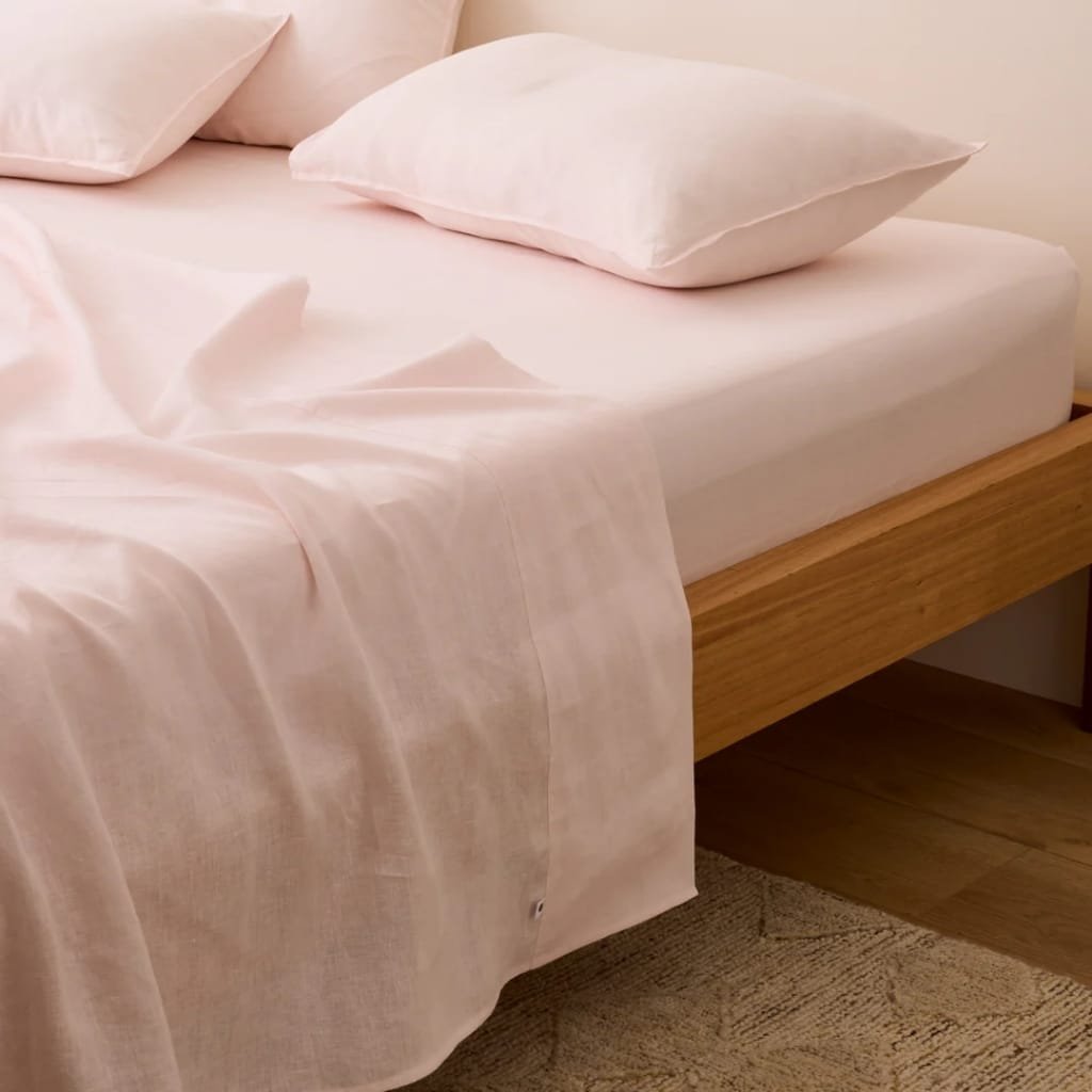 A neatly made bed with light pink sheets and matching pillowcases, placed on a wooden bed frame. Part of the light-colored carpet and cream-colored wall are visible.