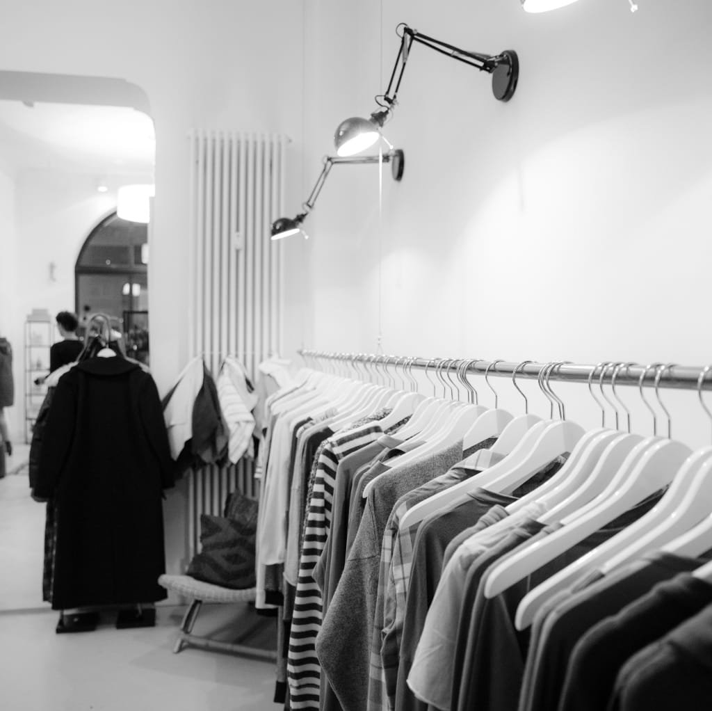 A black-and-white photo of a clothing rack filled with various T-shirts in a store or warehouse, with a person browsing in the background.
