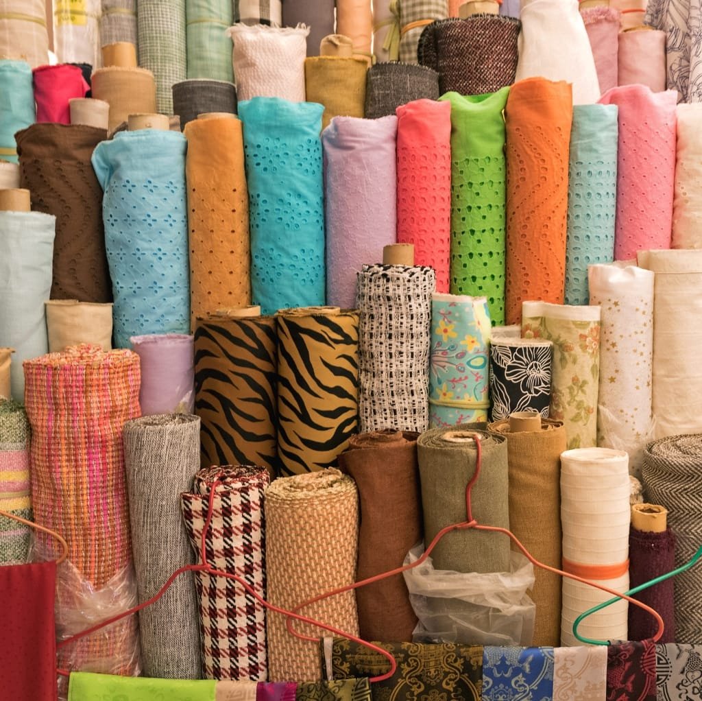  An assortment of colorful fabric rolls, primarily cotton, displayed at a wholesale fabric market, with various patterns and textures visible.