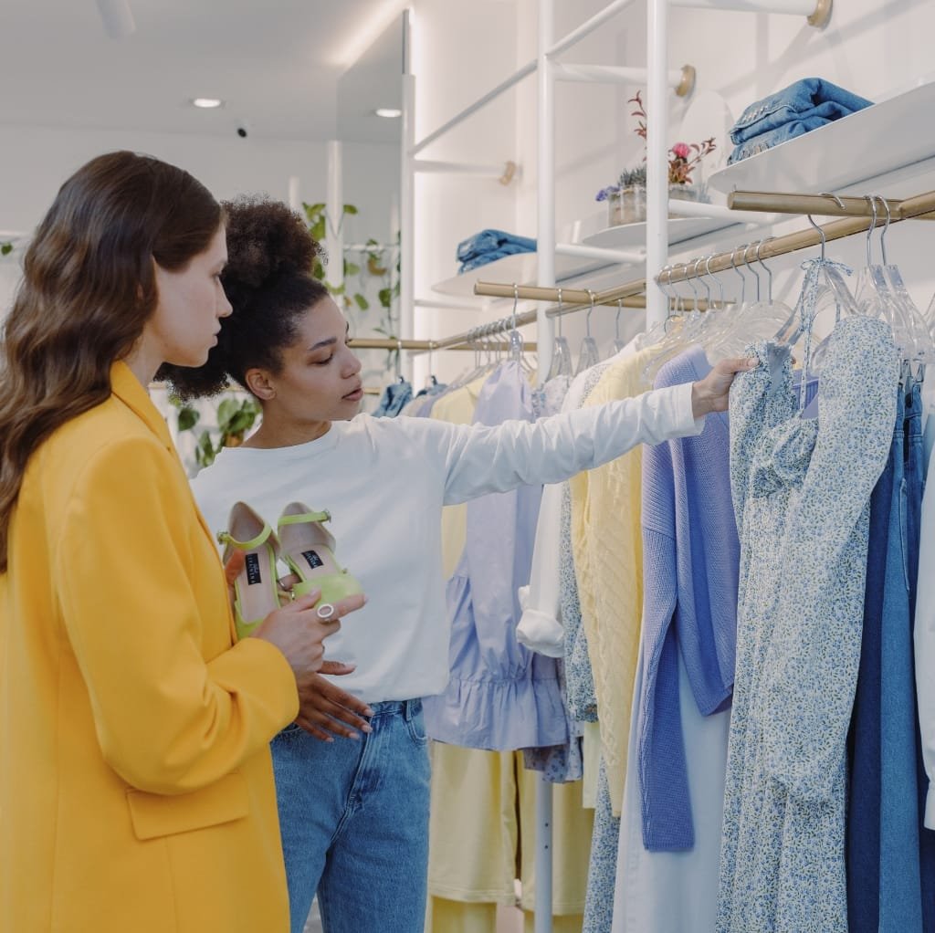 Two women shopping for cotton dresses in a clothing store, one holding a pair of shoes and pointing at a selection of light-colored dresses on a rack.