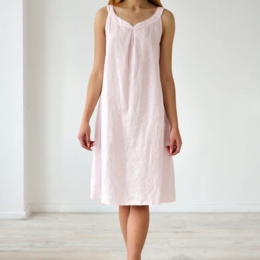 A woman wearing a light pink, sleeveless, knee-length nightgown with lace trim around the neckline, standing in a minimalistic room with light-colored wooden flooring and plain white walls.