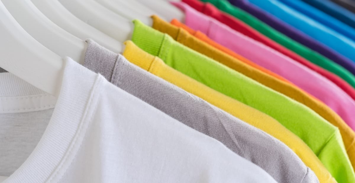 A row of colorful cotton t-shirts hanging on white hangers, showcasing various shades including white, gray, yellow, green, pink, purple, and blue.