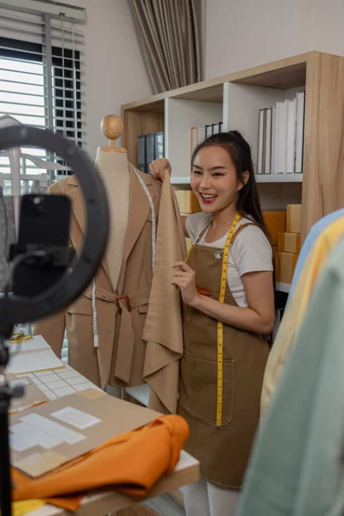 A young woman smiling and holding up a beige blazer on a mannequin while measuring it with a tape measure. She is in a workspace or studio with shelves, books, and fabric samples in the background. There is a camera on a tripod capturing the scene.