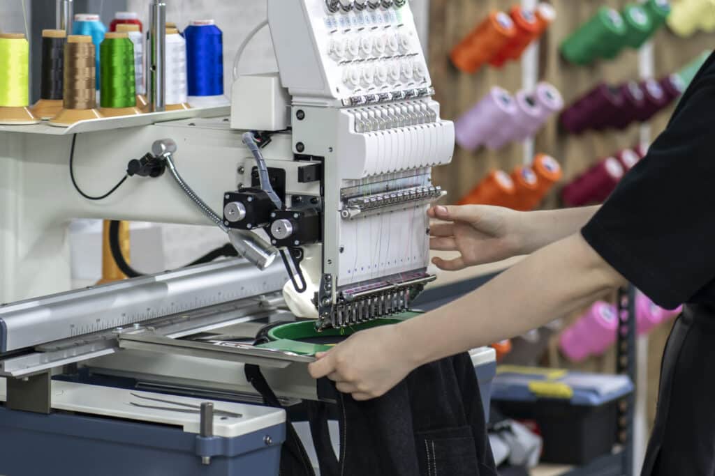 A person operating an industrial embroidery machine in a textile workshop, with colorful spools of thread in the background.