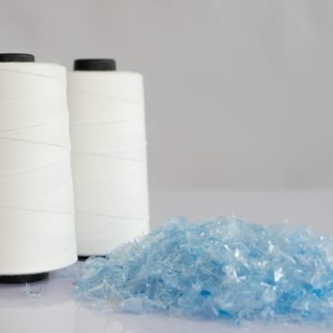 Spools of white thread with blue plastic flakes in front, illustrating the manufacturing process of PET polyester fabric.