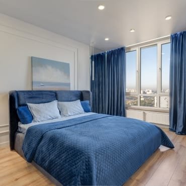 A modern bedroom featuring a bed with deep blue quilted bedding, matching pillows, large windows with blue curtains, and a cityscape view in the background.