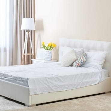 A cozy bedroom featuring a white bed with white bedding, decorative pillows, a side table with a vase of yellow flowers, a modern white floor lamp, and beige curtains.