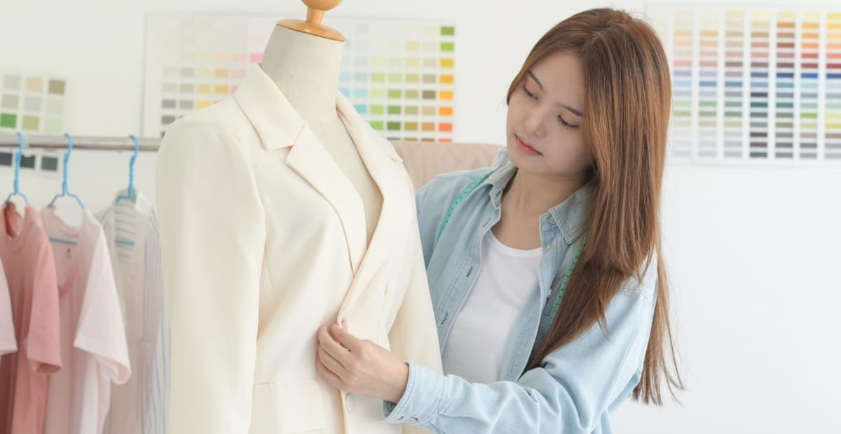 A woman examining a cream-colored blazer on a mannequin in a clothing store or design studio, with fabric swatches in the background.