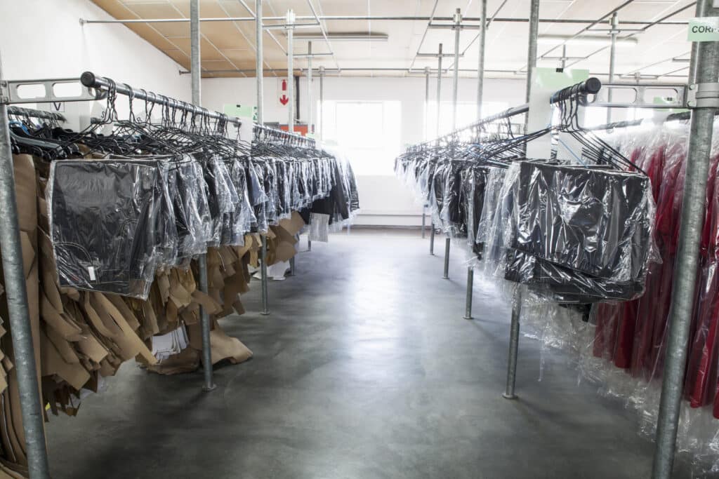 Rows of clothing on racks in a manufacturing or storage facility, with plastic-covered garments and paper pattern pieces, likely in a factory setting.