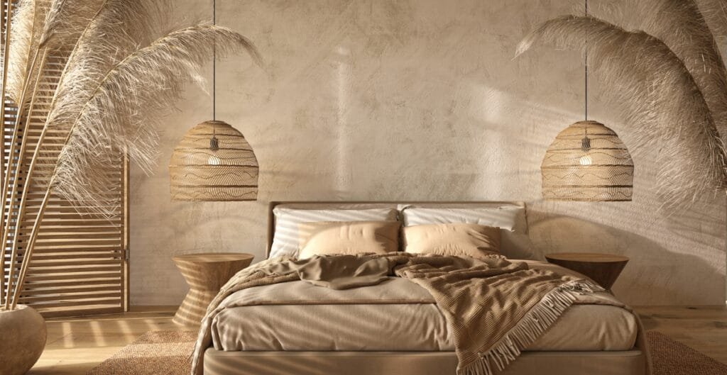 A cozy, warmly lit bedroom with a neutral color palette. The bed has beige bedding and pillows, with a blanket draped over the side. Two pendant lights with woven shades hang on either side of the bed. There are large, decorative pampas grass in a pot on the left, and the wall behind has a textured, rustic finish. A wooden slatted screen is visible in the corner.