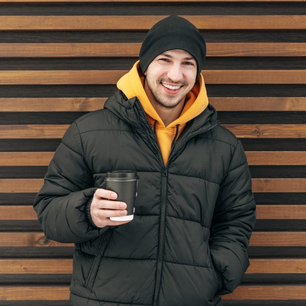 A smiling man dressed in a black 3-in-1 jacket, with a yellow hoodie underneath, holding a black travel mug and standing in front of a wooden wall.