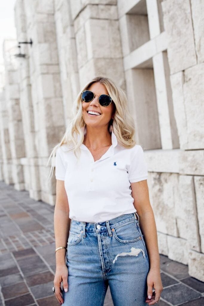  A woman smiling outdoors, wearing a white polo shirt and ripped blue jeans, accessorized with round sunglasses and bracelets, standing against a stone wall.

