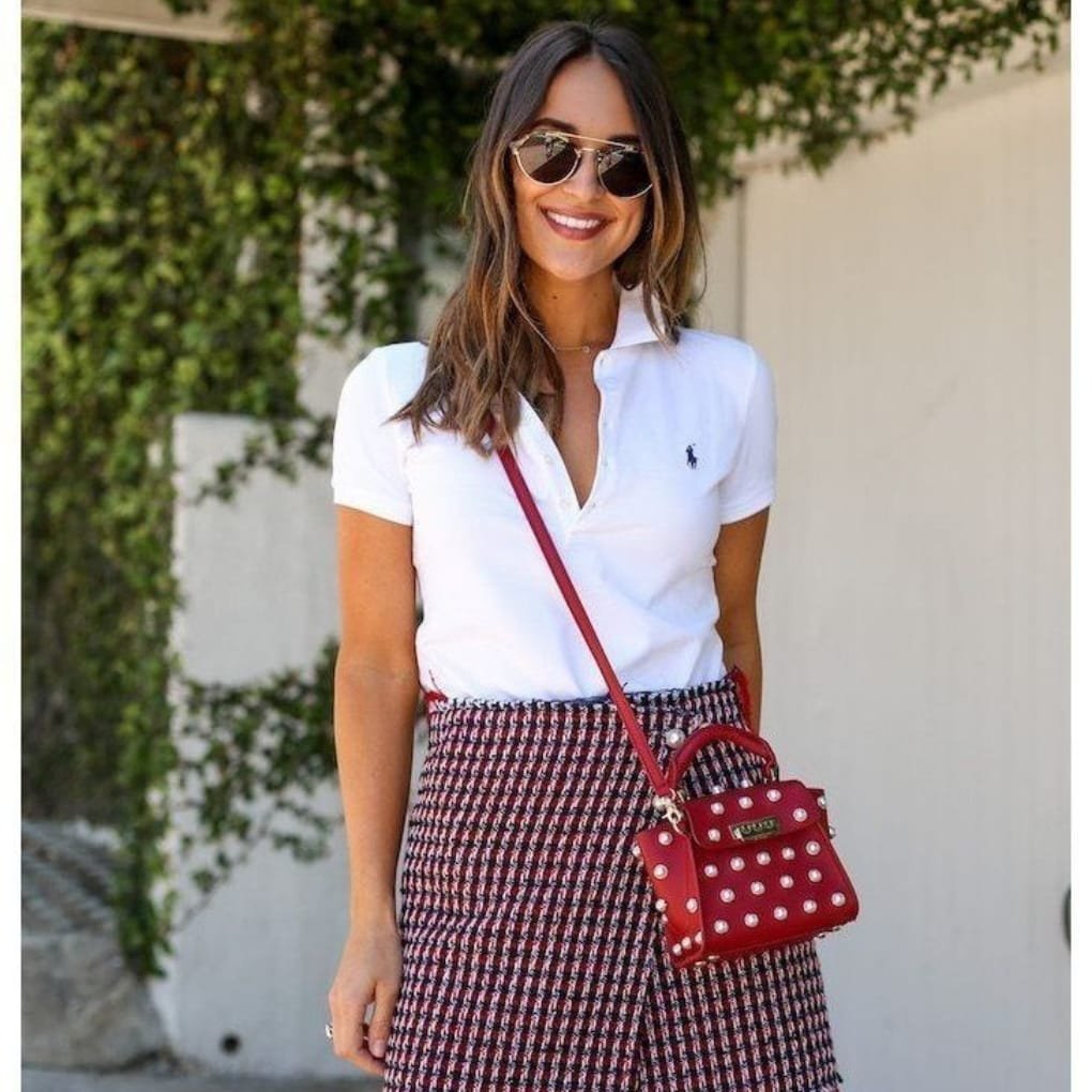 A woman smiling outdoors, wearing sunglasses, a white polo shirt, and a patterned skirt, carrying a red polka dot handbag.